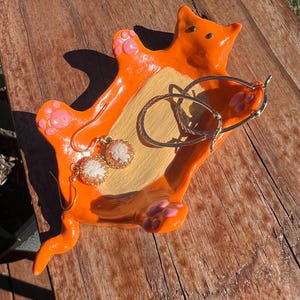 May include: Orange ceramic cat dish with pink paw prints, holding gold hoop earrings and small white cameo earrings.