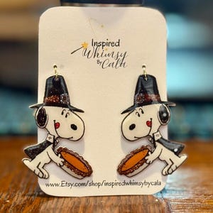 May include: A pair of dangle earrings featuring a cartoon dog character wearing a black hat and cape, holding a hot dog. The earrings are white, black, and brown with gold-colored earring hooks. The text "Inspired Whimsy by Cala" and a website address are visible.