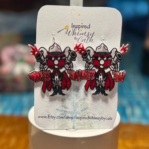 May include: A pair of novelty earrings featuring a cartoon character in a cowboy hat, holding two guns. The character is wearing a red cape and the text "Wreck 'Em!" is displayed. The earrings are on a white card.