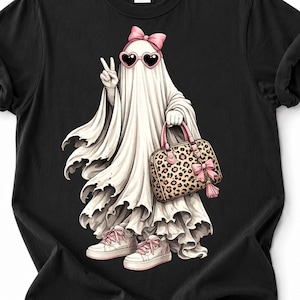 Peut inclure: Un t-shirt noir avec un motif représentant un fantôme blanc portant un nœud rose, des lunettes de soleil en forme de cœur et des baskets. Le fantôme fait un signe de paix et porte un sac à main à motif léopard avec un nœud et un pompon roses.