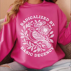 Pu&ograve; includere: Una felpa magenta con la scritta "RADICALIZED BY BASIC DECENCY" in un disegno floreale bianco con un uccello. La felpa ha maniche lunghe e girocollo. Il disegno &egrave; centrato sul davanti.
