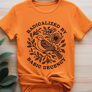 Pu&ograve; includere: T-shirt arancione con una grafica nera di un uccello posato su un ramo, circondato da foglie e fiori. Il testo "RADICALIZED BY BASIC DECENCY" &egrave; disposto a cerchio attorno al disegno dell'uccello.