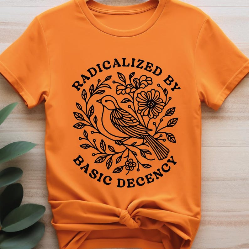 Pu&ograve; includere: T-shirt arancione con una grafica nera di un uccello posato su un ramo, circondato da foglie e fiori. Il testo "RADICALIZED BY BASIC DECENCY" &egrave; disposto a cerchio attorno al disegno dell'uccello.