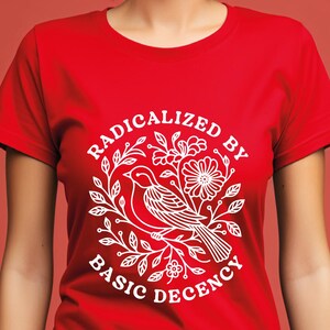 Pu&ograve; includere: T-shirt rossa con grafica bianca. La grafica raffigura un uccello su un ramo fiorito, circondato dalla scritta "RADICALIZED BY BASIC DECENCY". Il disegno &egrave; centrato sulla parte anteriore della maglietta.