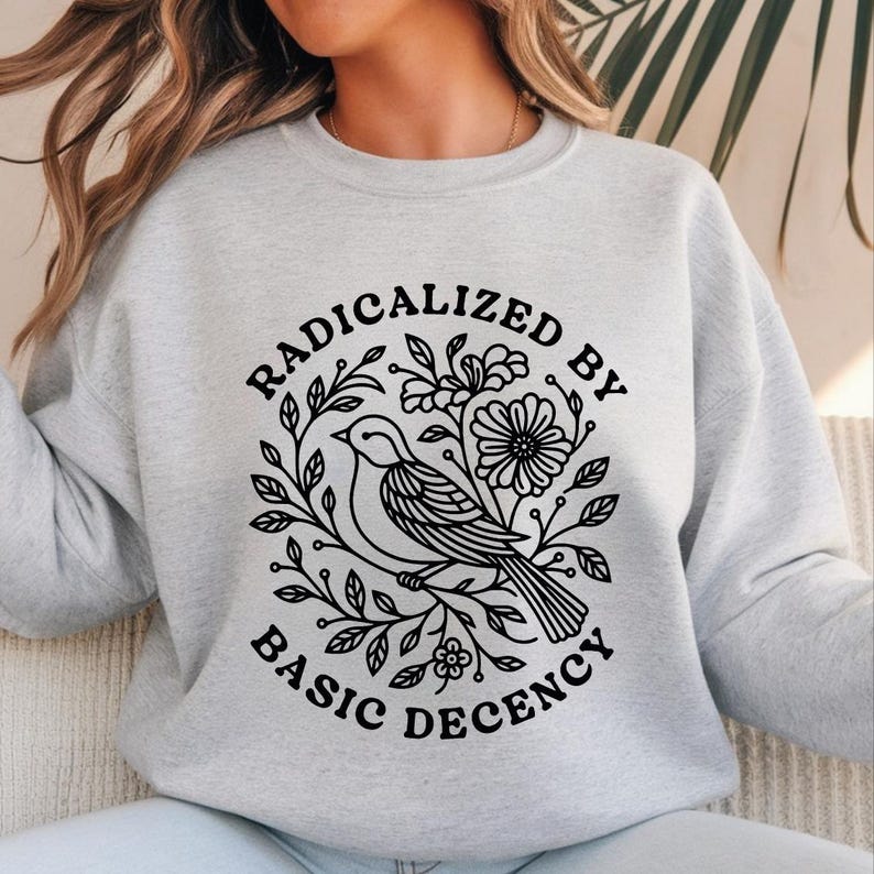 Pu&ograve; includere: Felpa grigio chiaro con grafica nera. La grafica presenta un uccello posato su un ramo, circondato da foglie e fiori, con le parole "RADICALIZED BY BASIC DECENCY" in un design circolare.
