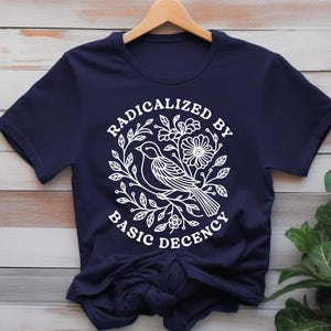 Pu&ograve; includere: T-shirt blu navy con grafica bianca. La grafica presenta un uccello, fiori e la scritta "RADICALIZED BY BASIC DECENCY" in un cerchio. La maglietta &egrave; appesa a una gruccia di legno.
