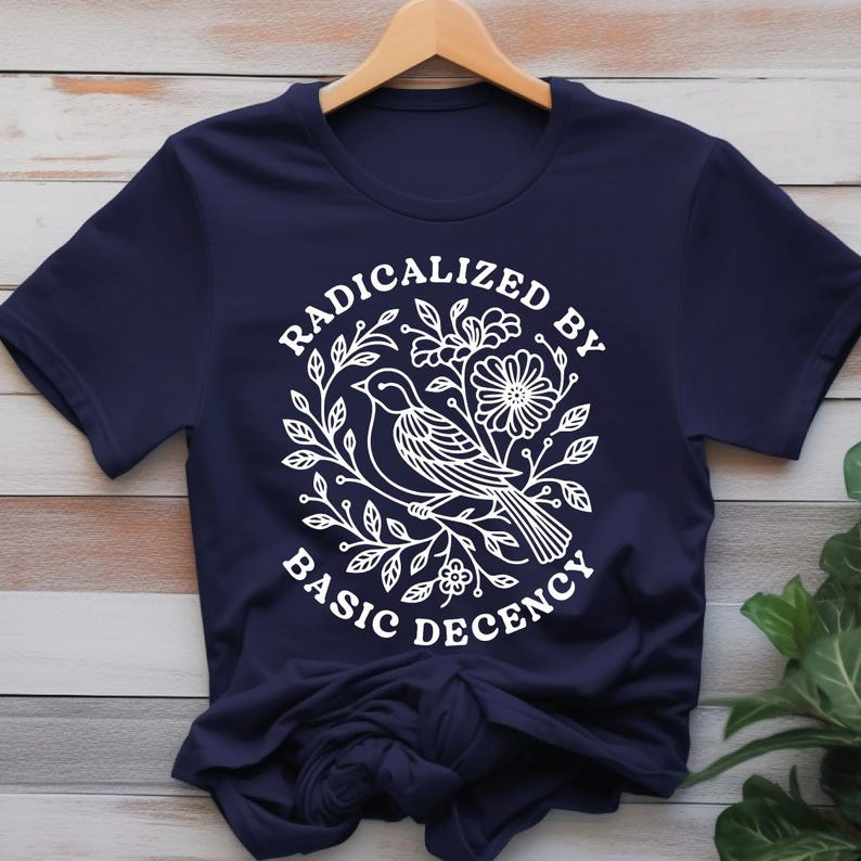 Pu&ograve; includere: T-shirt blu navy con grafica bianca. La grafica presenta un uccello, fiori e la scritta "RADICALIZED BY BASIC DECENCY" in un cerchio. La maglietta &egrave; appesa a una gruccia di legno.