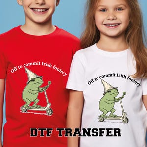 May include: Red and white t-shirts with a graphic of a green frog on a scooter, wearing a leprechaun hat and holding a four-leaf clover. The text "Off to commit Irish foolery" is above the frog. The text "DTF TRANSFER" is at the bottom.