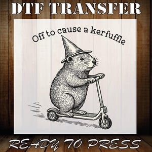 May include: DTF transfer design featuring a detailed illustration of a rodent wearing a witch hat and riding a scooter. The text "Off to cause a kerfuffle" arches above the rodent, with "READY TO PRESS" below.