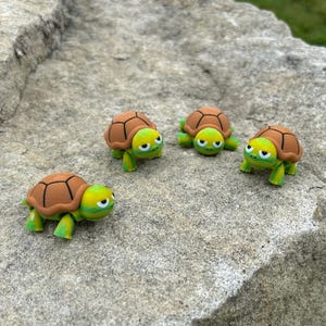 May include: Four small, cartoon-style turtle figurines. Each turtle has a brown shell, green legs, and a yellow and green head. The turtles are arranged on a gray, textured rock surface.