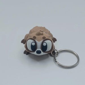 May include: A brown and tan meerkat keychain with large, expressive eyes and a silver keyring. The meerkat has a textured body and is lying down. The keychain is a fun accessory.