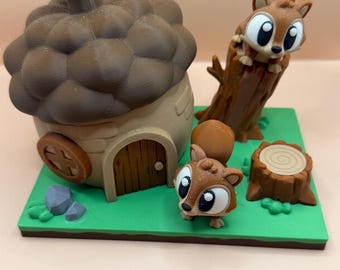 May include: A whimsical diorama featuring a brown acorn-shaped house with a door and a round window. Two cartoon squirrels are present, one peeking from a tree stump and another standing on a green base. The scene includes a tree stump and two gray rocks.