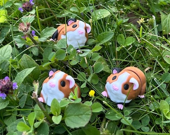 May include: Three small, handcrafted guinea pig figurines with brown, white, and black markings are nestled in green grass and clover. The guinea pigs have pink ears and feet. Purple and yellow wildflowers are also visible.