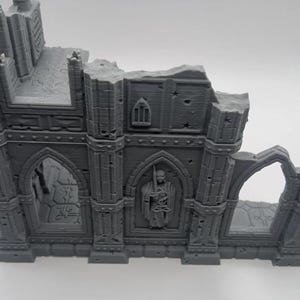 May include: A gray 3D printed model of a ruined building with a gothic archway and a figure carved into the wall. The model is detailed with stonework and has a weathered look.
