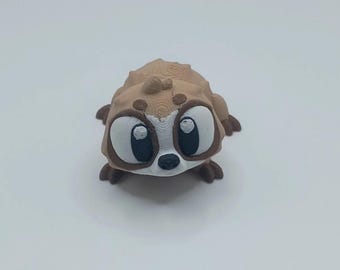 May include: A 3D-printed figurine of a cartoon meerkat. The meerkat has large, expressive eyes, a small nose, and a textured body in shades of brown and tan. The figurine is designed to sit upright.