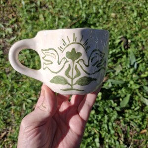 May include: White ceramic mug with a green hand-painted design of two birds facing each other with a green plant and sun rays in the center.