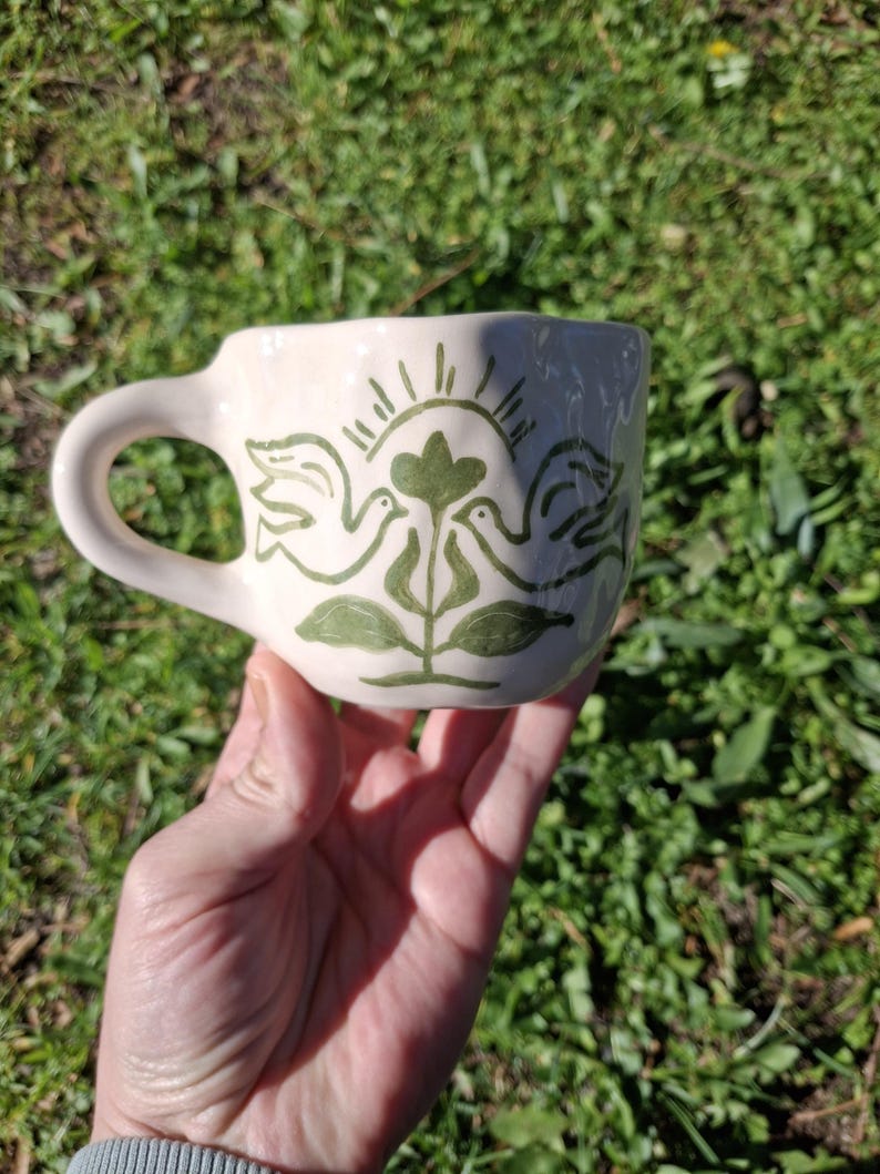 May include: White ceramic mug with a green hand-painted design of two birds facing each other with a green plant and sun rays in the center.