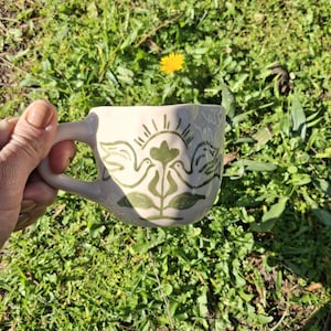 May include: A white ceramic mug with a green design of two birds facing each other with a flower in the middle. The birds have wings and a sun with rays behind them.