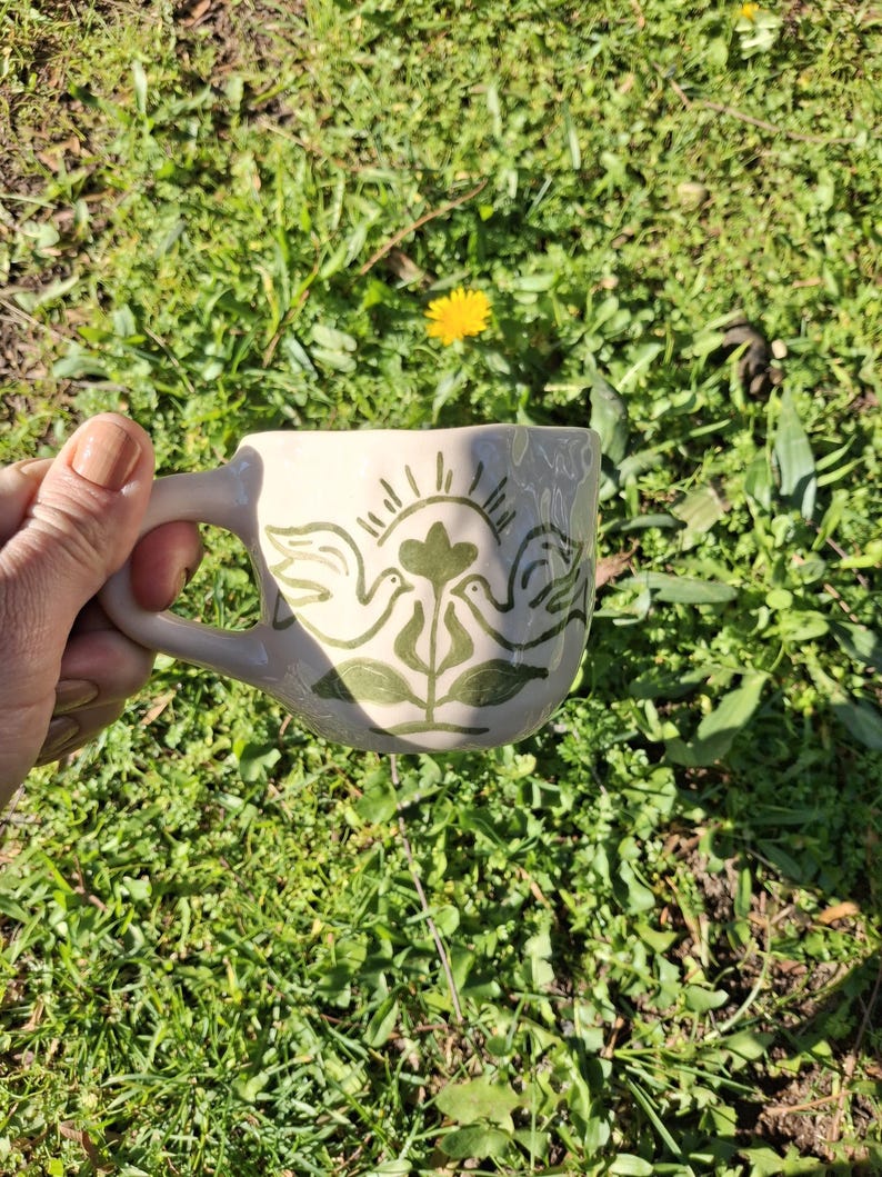 May include: A white ceramic mug with a green design of two birds facing each other with a flower in the middle. The birds have wings and a sun with rays behind them.