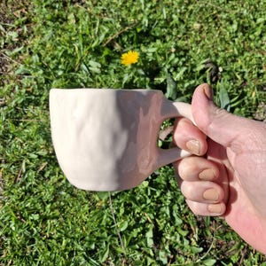 May include: A white ceramic mug with a textured surface and a handle. The mug is being held in a hand.