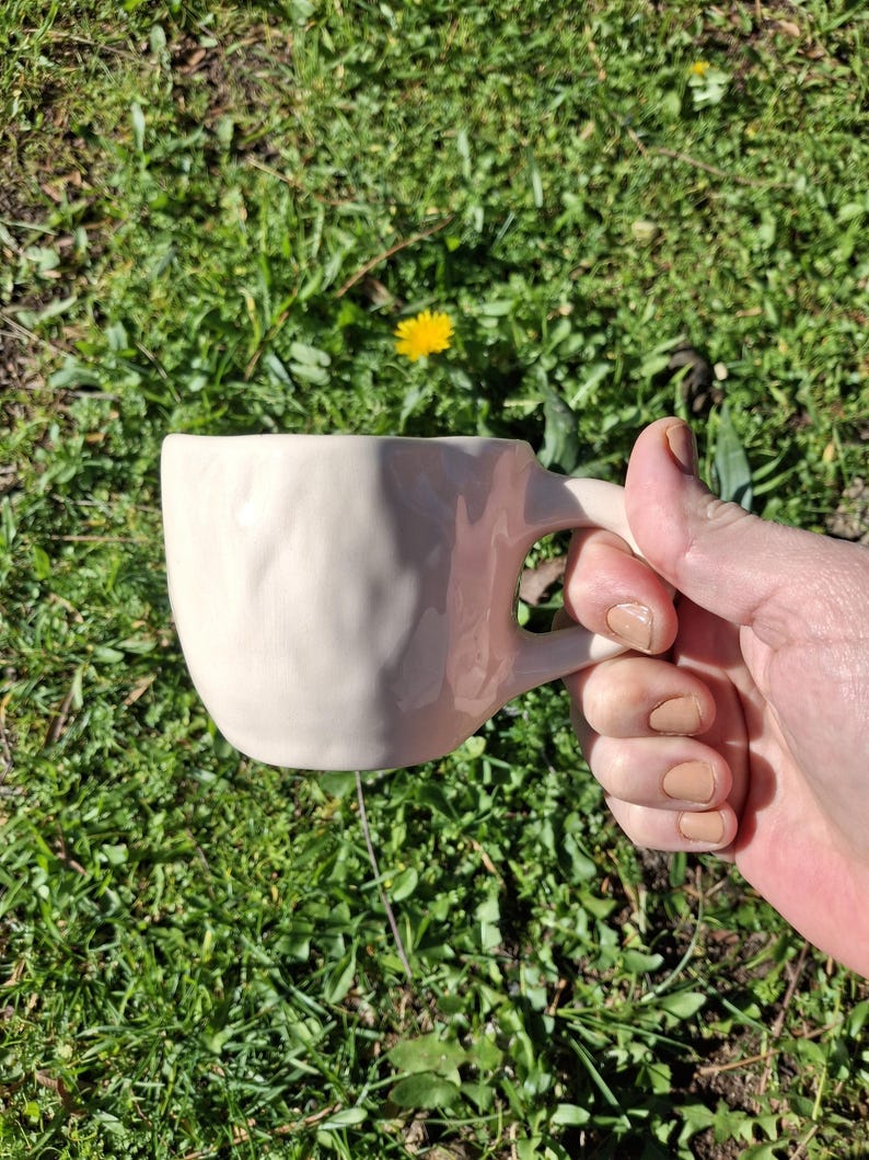 May include: A white ceramic mug with a textured surface and a handle. The mug is being held in a hand.