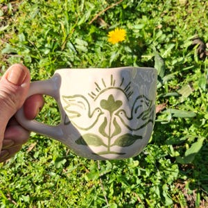 May include: A white ceramic mug with a green design of two birds and a flower. The design is reminiscent of folk art.
