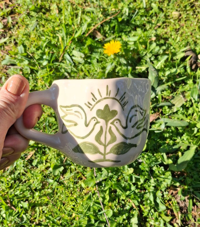 May include: A white ceramic mug with a green design of two birds and a flower. The design is reminiscent of folk art.