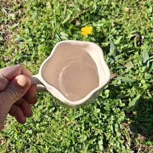 May include: A white ceramic mug with a wavy, uneven rim and a handle. The mug is empty and is being held by a hand against a green grassy background.