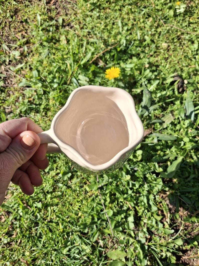 May include: A white ceramic mug with a wavy, uneven rim and a handle. The mug is empty and is being held by a hand against a green grassy background.