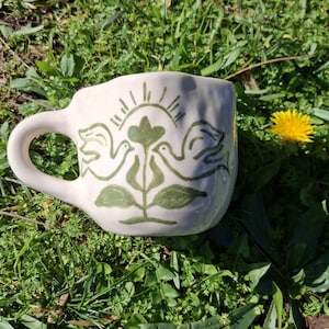 May include: A white ceramic mug with a green hand-painted design of two birds perched on a plant with a sun above them.
