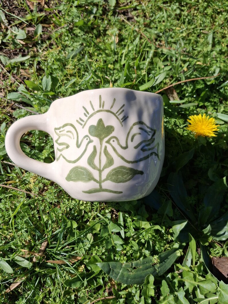 May include: A white ceramic mug with a green hand-painted design of two birds perched on a plant with a sun above them.