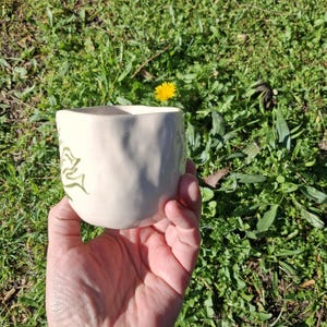May include: A white ceramic mug with a green leafy design. The mug has a textured surface and is being held in a hand.