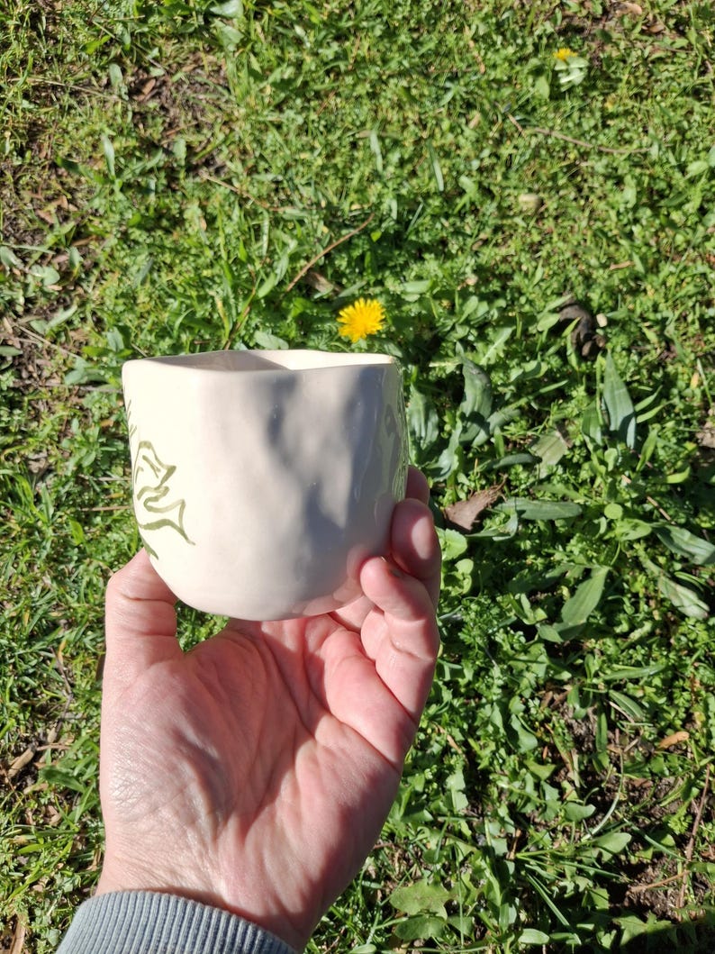 May include: A white ceramic mug with a green leafy design. The mug has a textured surface and is being held in a hand.