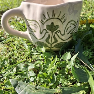 May include: A white ceramic mug with a green design of two birds and a heart. The mug has a wavy rim and a handle.