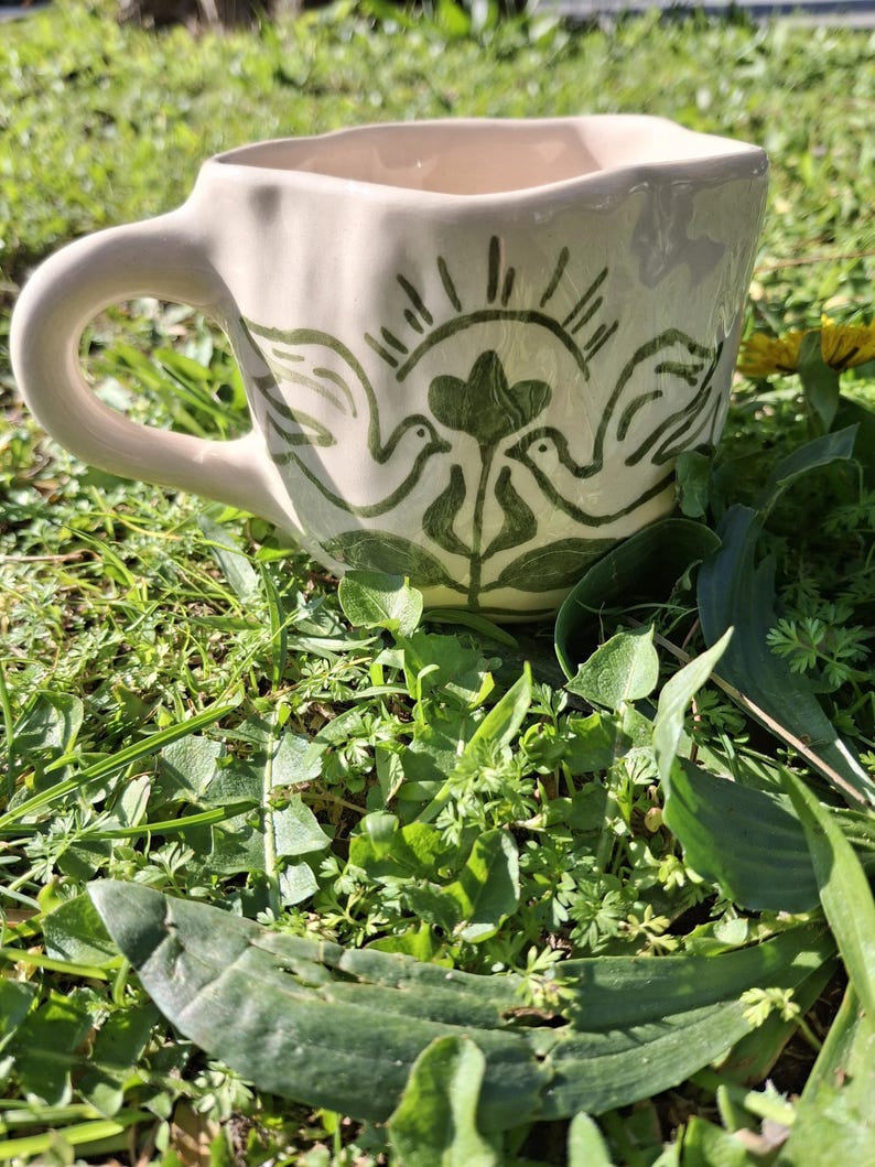 May include: A white ceramic mug with a green design of two birds and a heart. The mug has a wavy rim and a handle.