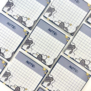 May include: Square notepads with a light blue border and grid pattern. Each notepad has the word "NOTE:" printed at the top and a decorative illustration of a whale and stars in the corner. The notepads are arranged on a white surface.