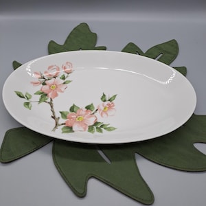 Könnte beinhalten: Ovale weiße Servierplatte mit einem Blumendesign aus rosa Blüten und grünen Blättern. Die Platte liegt auf einem grünen, blattförmigen Platzset. Ein dekoratives Stück zum Servieren von Speisen.