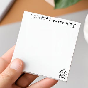 以下が含まれることがあります： 「I ChatGPT everything!」と黒字で書かれた白い正方形の付箋。小さな笑顔のロボットのイラストが右下にあります。メモは手で持たれています。