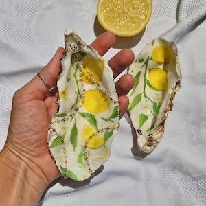 Puede incluir: Dos conchas de ostra pintadas con un diseño de limón. Una concha se sostiene en una mano y la otra está al lado. Una mitad de limón está en el fondo. Las conchas están pintadas con limones amarillos y hojas verdes.