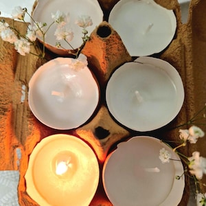 May include: Five white tea lights are placed in an egg carton. One tea light is lit and glowing orange. The carton is brown and has a rough texture.