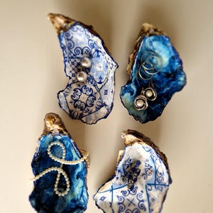 Könnte beinhalten: Vier blaue und weiße Austernmuscheln mit dekorativen Designs. Jede Schale enthält Schmuck, darunter eine Perlenkette, Ohrringe und einen Ring.