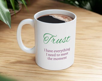 Puede incluir: Una taza de cerámica blanca con la palabra Trust en letra cursiva verde, seguida del texto I have everything I need to meet the moment en una fuente sans-serif de color morado. La taza está llena de un líquido oscuro, como café o té.