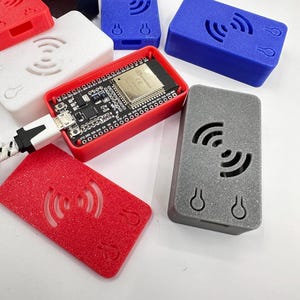 Könnte beinhalten: Eine Sammlung kleiner, rechteckiger elektronischer Geräte in verschiedenen Farben, darunter Rot, Blau, Weiß und Grau. Einige Geräte haben eine sichtbare Leiterplatte, die über ein USB-Kabel verbunden ist. Die Geräte verfügen über ein drahtloses Signalsymbol und ein schlüssellochförmiges Design.
