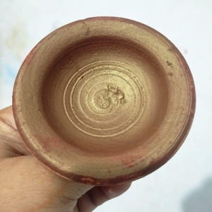 May include: A small, round, terracotta-colored bowl with a gold-painted interior. The bowl's rim is painted red. The interior features concentric circles and a textured design at the center. The bowl is held by a hand.