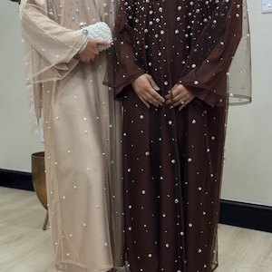May include: Two floor-length abayas, one in beige and one in dark brown, both featuring sheer overlay fabric embellished with scattered silver-toned beads and crystals. Each garment is paired with matching inner layers and pointed-toe heels.