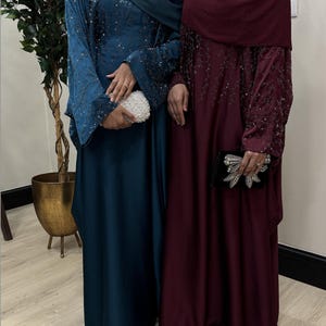 May include: Two floor-length abayas, one in deep teal and one in burgundy, both featuring long sleeves with intricate beadwork and sequin embellishments. The teal abaya is paired with a matching headscarf and a white beaded clutch, while the burgundy abaya includes a matching headscarf and a black clutch with a large decorative bow.
