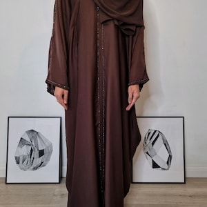 May include: A long, flowing brown abaya with embellished trim and sleeves. The garment is worn with a matching head covering. Two framed black and white geometric art prints are visible in the background.
