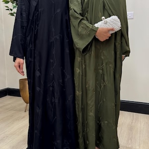 Könnte beinhalten: Zwei bodenlange Abayas, eine in einfarbigem Schwarz und eine in Olivgrün, beide mit langen Ärmeln und einem zarten, tonalen Ranken- und Blattstickmuster. Die olivgrüne Abaya wird mit einer weißen, perlenbesetzten Clutch mit silbernem Verschluss kombiniert.