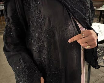May include: Black abaya with intricate floral embroidery and sparkling embellishments. The sheer fabric reveals a glimpse of a patterned underdress. A black handbag with a crocodile texture is held in the hand.