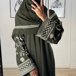 Peut inclure: Une abaya vert olive avec des broderies complexes sur les manches et l'encolure. Les manches présentent des motifs floraux et géométriques en fil d'or et blanc. Le vêtement est une conception fluide et modeste.